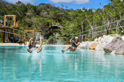 Dos visitantes disfrutan de un emocionante paseo en tirolesa que termina con un chapuzón en una piscina de aguas cristalinas en Scape Park Cap Cana.
