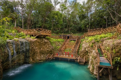 Un cenote de agua azul profunda rodeado de acantilados rocosos, árboles verdes y escaleras de madera que bajan al agua.